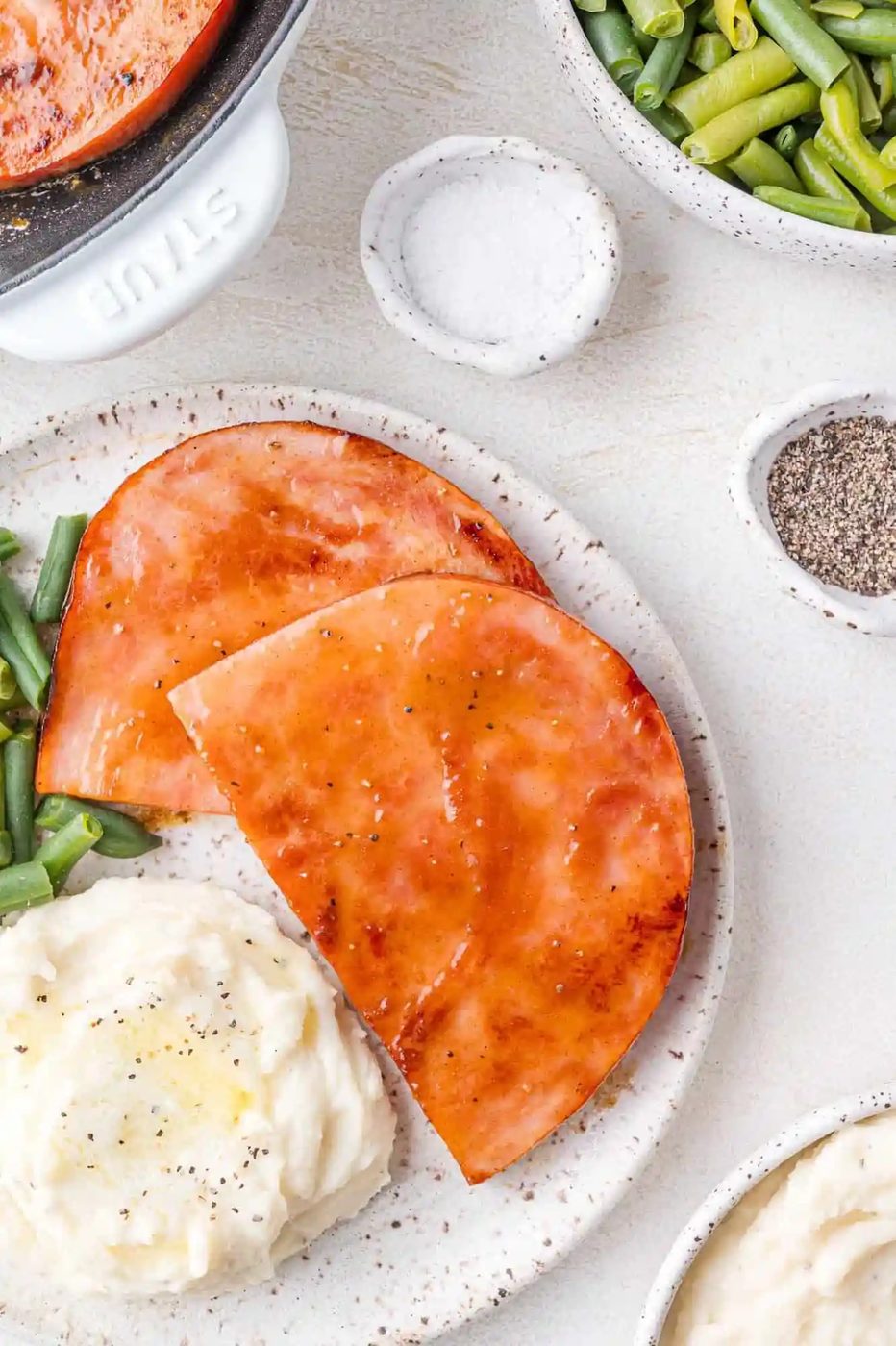 Plate with ham slices, mashed potatoes topped with butter, and green beans, surrounded by small bowls of salt and pepper on a white surface.