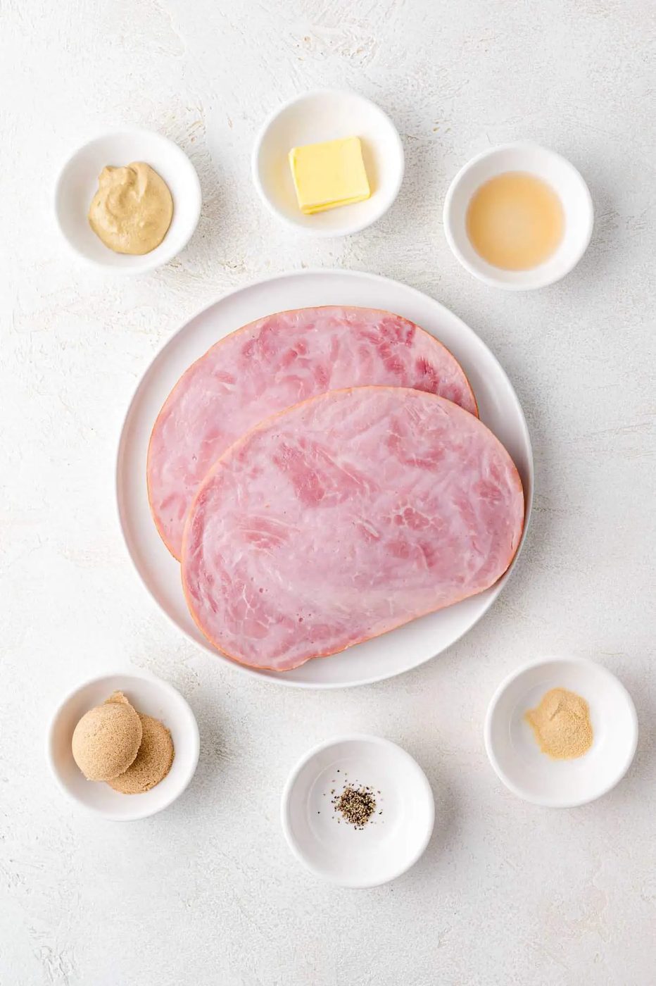 Plate with two slices of ham surrounded by small bowls containing mustard, butter, vinegar, brown sugar, black pepper, and powdered spice on a light textured surface.