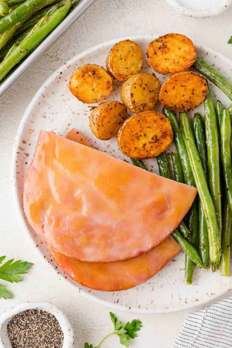 A plate with slices of pink ham, seasoned roasted potato halves, and green beans next to it. Plate is set on a textured white surface.