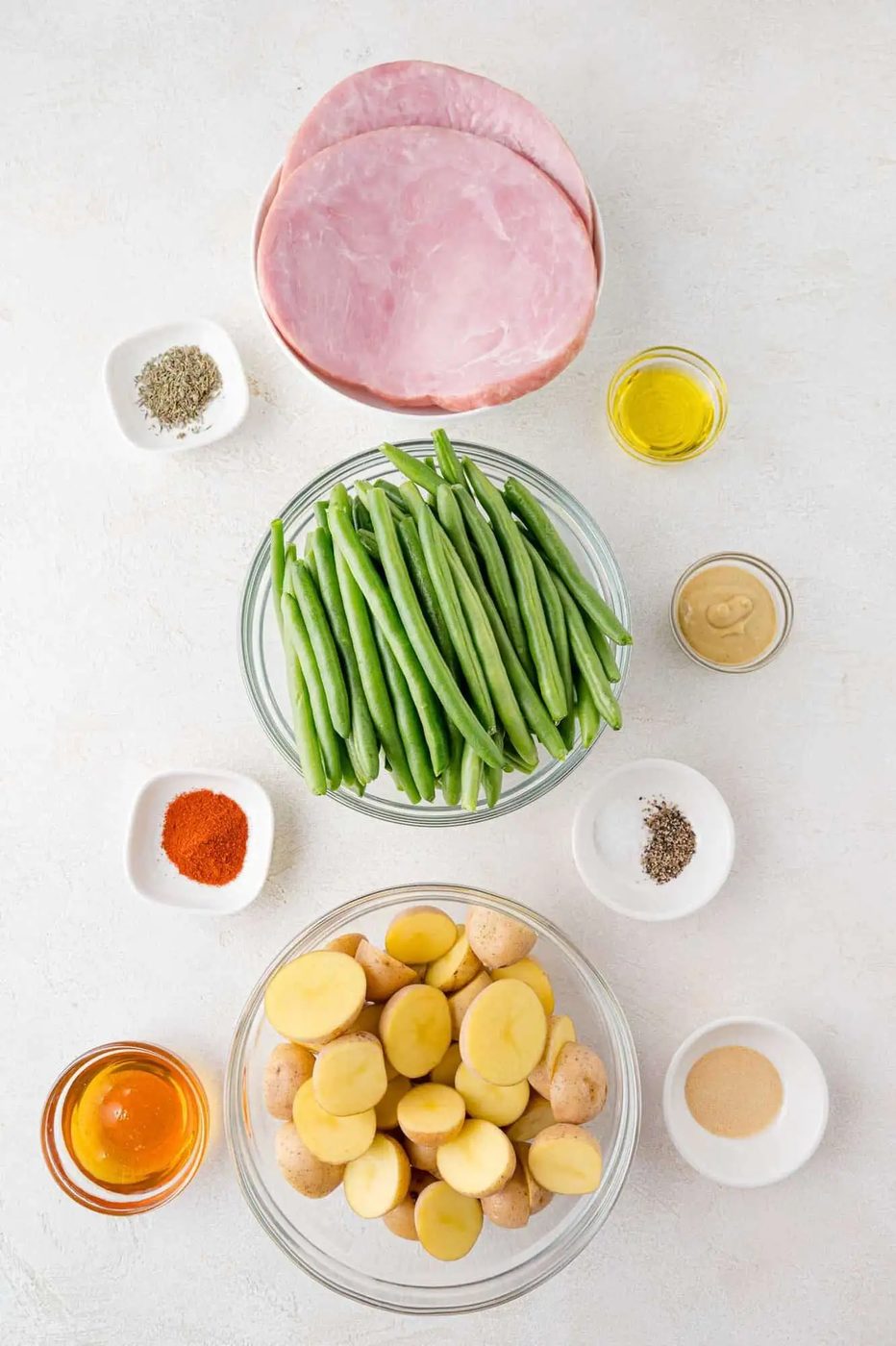 Ingredients arranged on a surface: slices of ham, fresh green beans, halved baby potatoes, olive oil, mustard, seasonings, paprika, honey, and spices in small bowls.