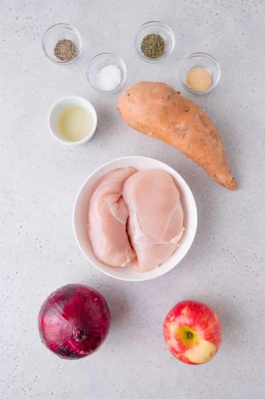 Raw chicken breasts in a bowl, sweet potato, red onion, apple, oil, salt, pepper, and garlic powder arranged on a light gray surface.