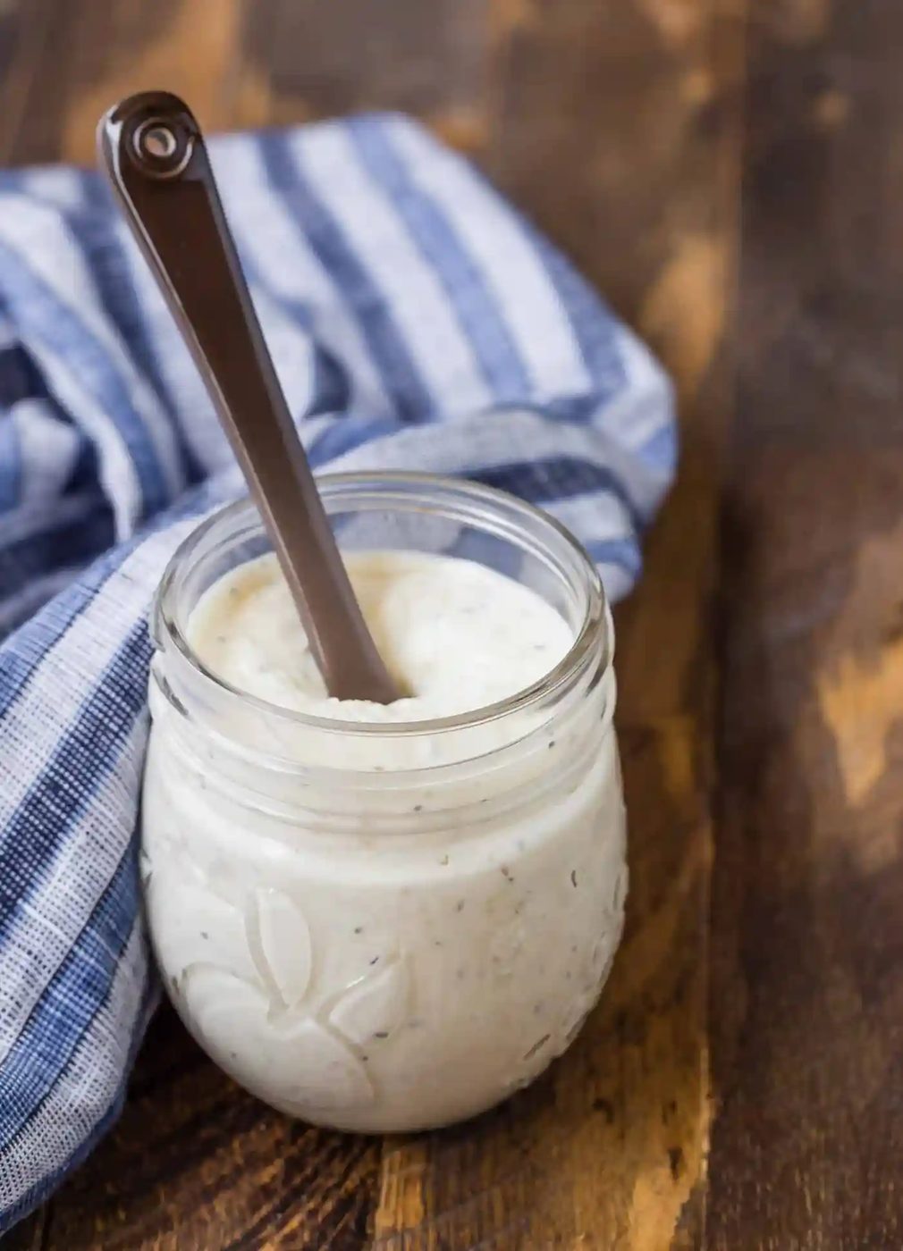 Small glass jar of a creamy caesar dressing on a wooden background. A blue and white linen is also pictured, and a small brown glass spoon.