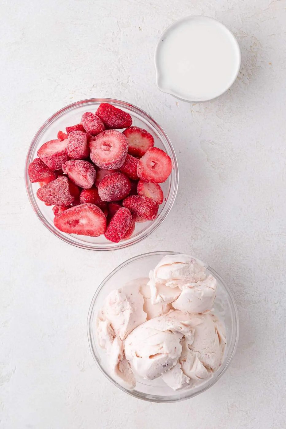 A bowl of frozen strawberries, a bowl of strawberry ice cream, and a small pitcher of milk on a white surface.