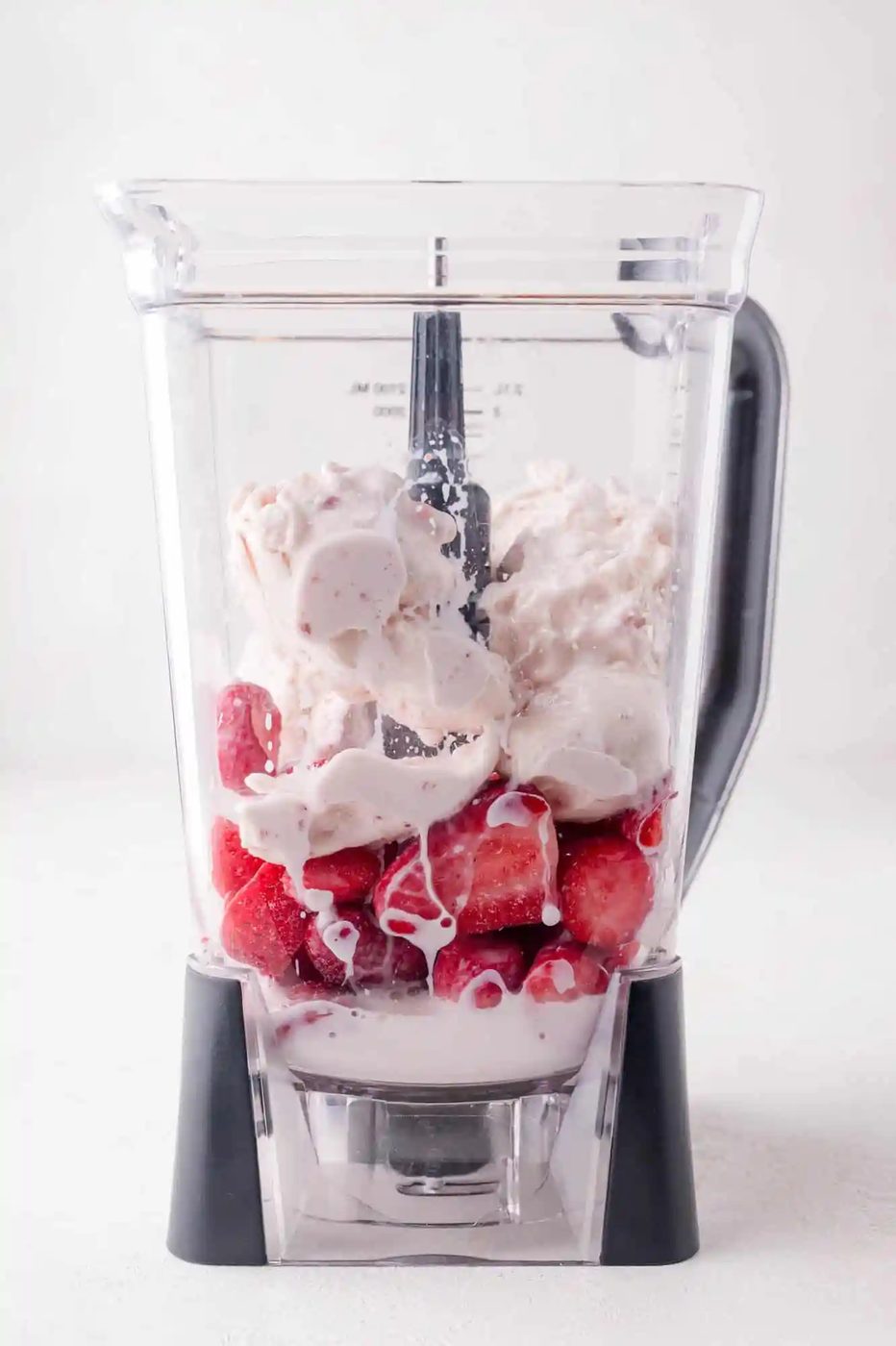 A blender filled with frozen strawberries, dollops of vanilla ice cream, and a small amount of milk, ready to be blended.