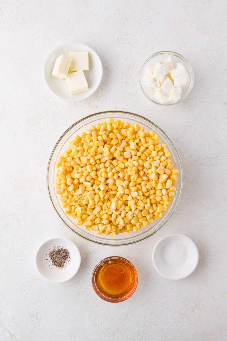 A bowl of corn kernels surrounded by small bowls of butter, sour cream, salt, pepper, and honey on a white surface.