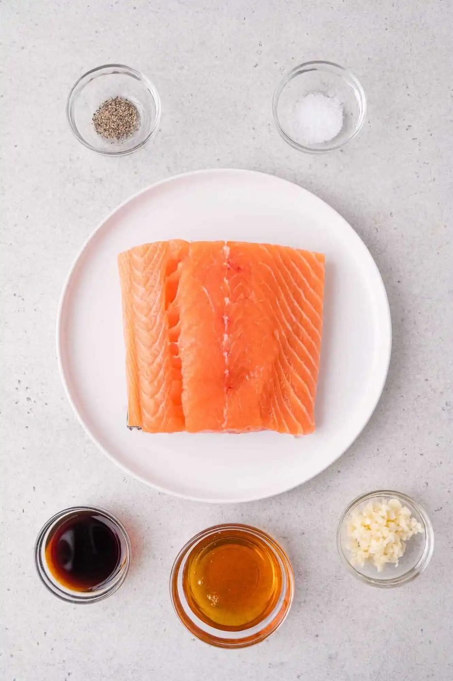 Raw salmon fillets on a white plate, surrounded by bowls of soy sauce, honey, minced garlic, black pepper, and salt on a light gray surface.