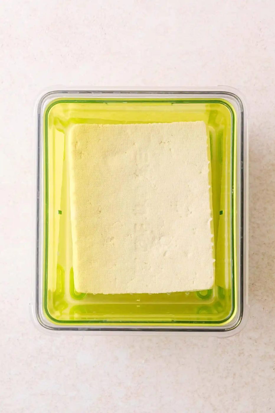 A block of tofu sits inside a green plastic container, viewed from above on a light-colored surface.