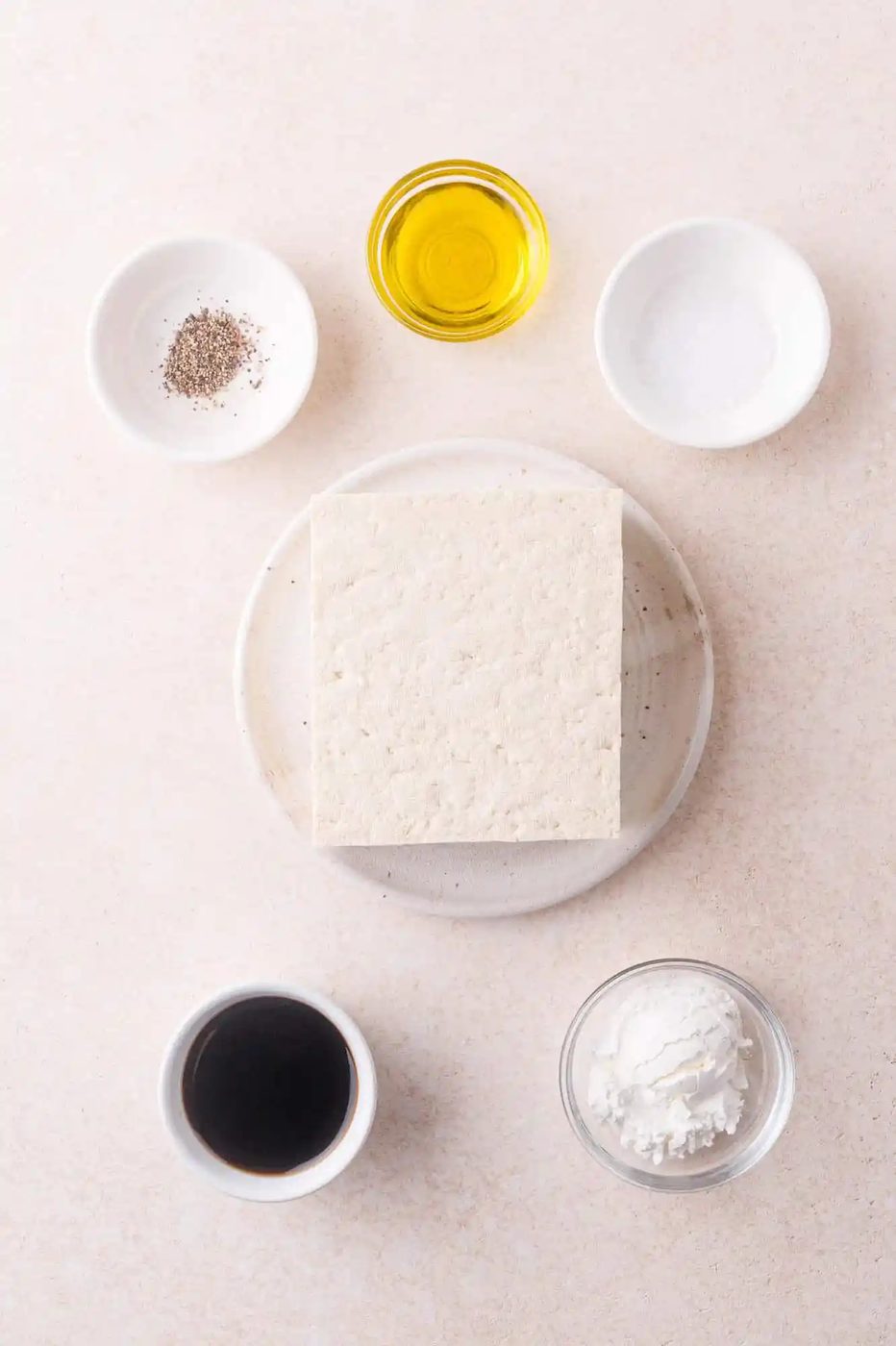 A block of tofu on a plate surrounded by small bowls containing black pepper, olive oil, salt, soy sauce, and cornstarch on a light surface.