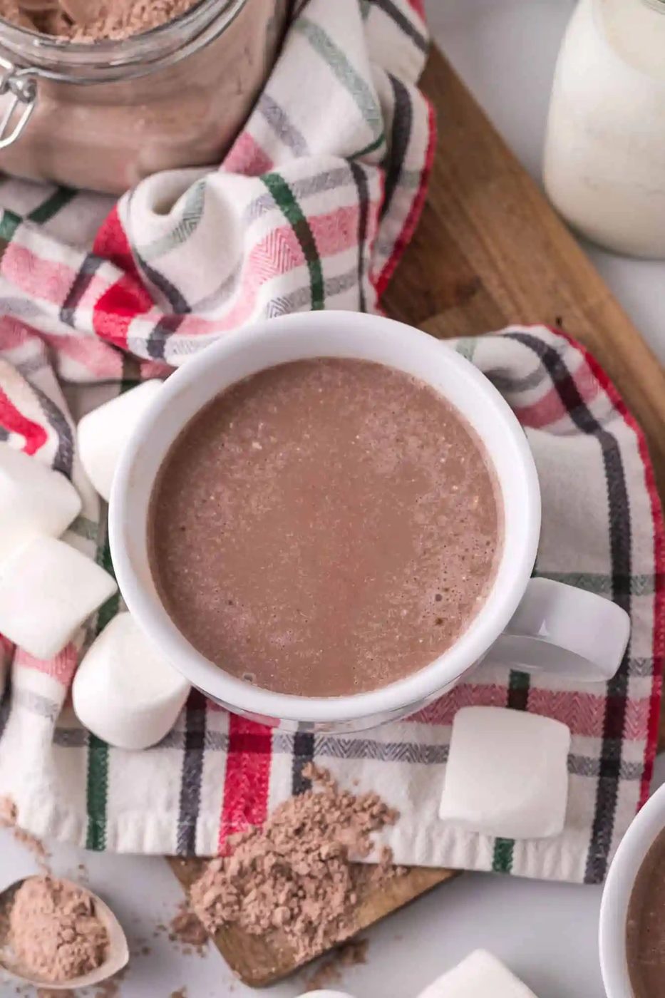 A mug of hot chocolate on a plaid cloth, surrounded by marshmallows and cocoa powder.