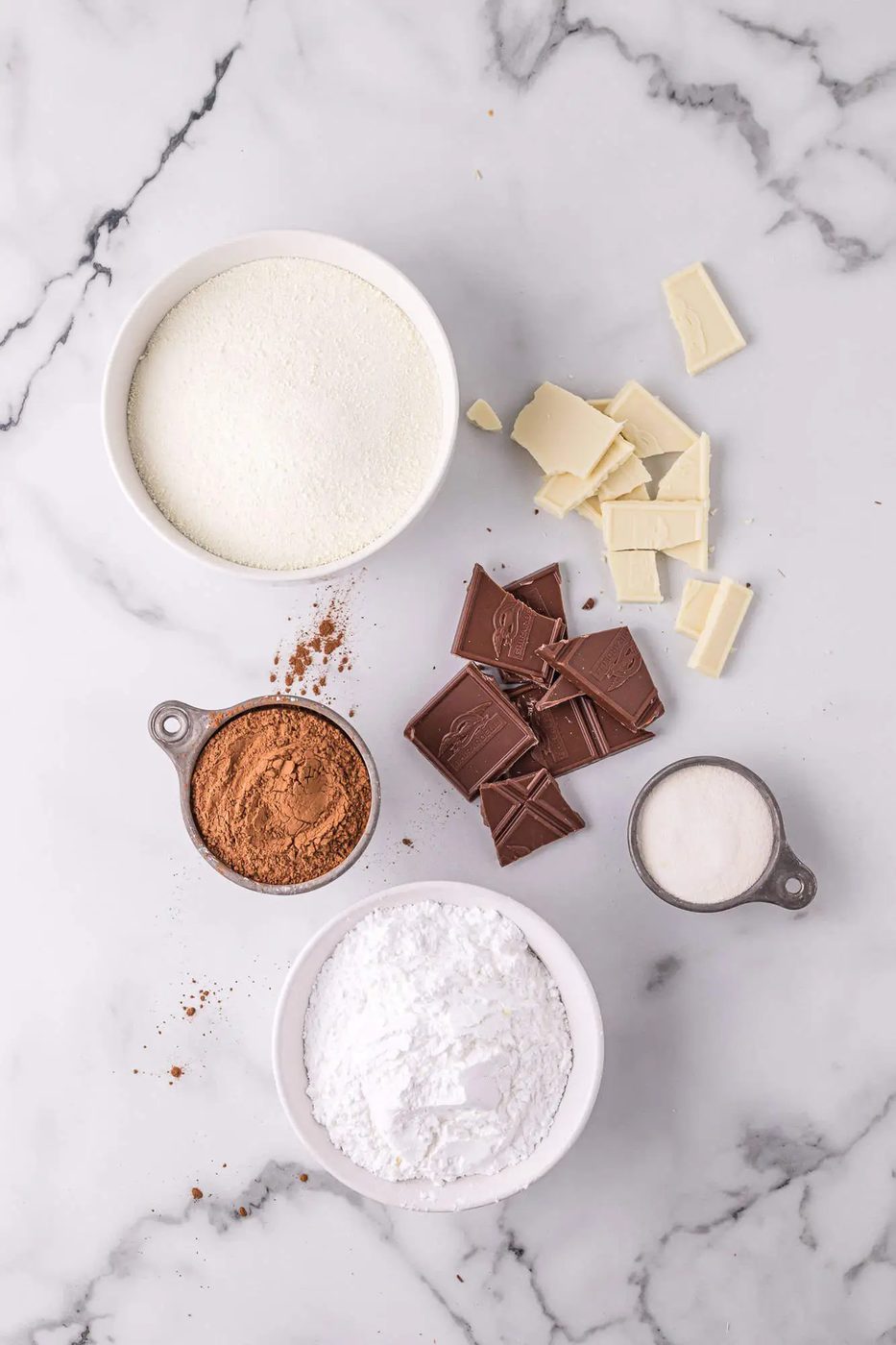 Ingredients on a marble surface: bowls of sugar, cocoa powder, and sugar, with pieces of white and dark chocolate.