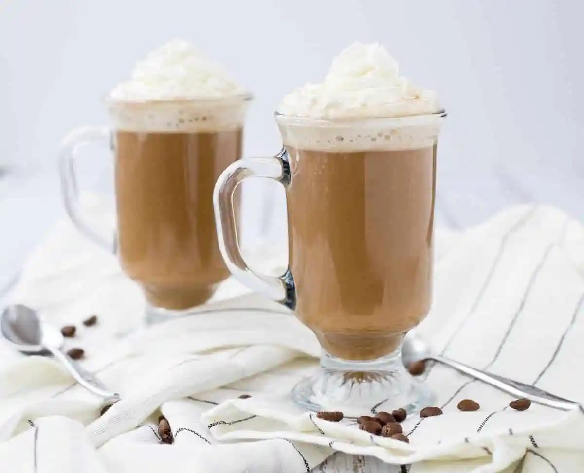 Front view of two mugs of white Russian hot chocolate with two spoons on cloth.