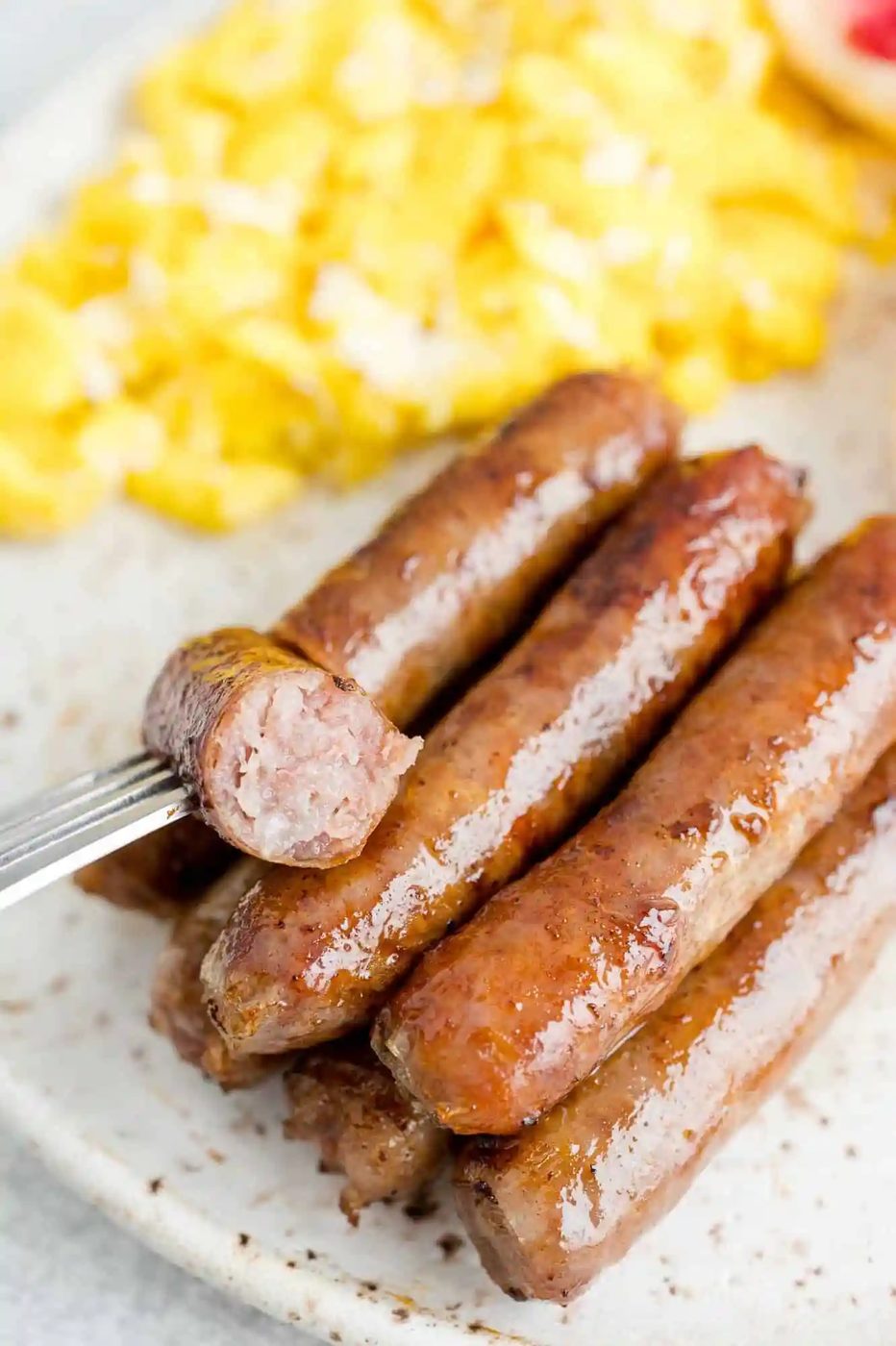 A plate with cooked breakfast sausages and scrambled eggs, with a fork holding a cut piece of sausage.