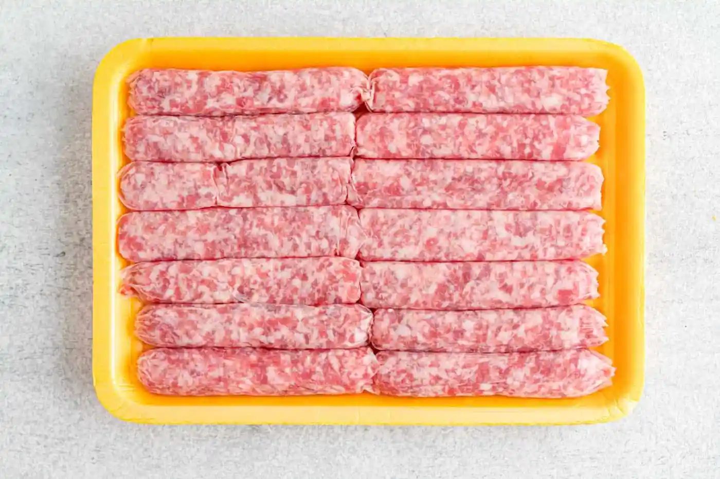 A yellow tray with raw sausage links arranged in two neat rows on a light-colored surface.