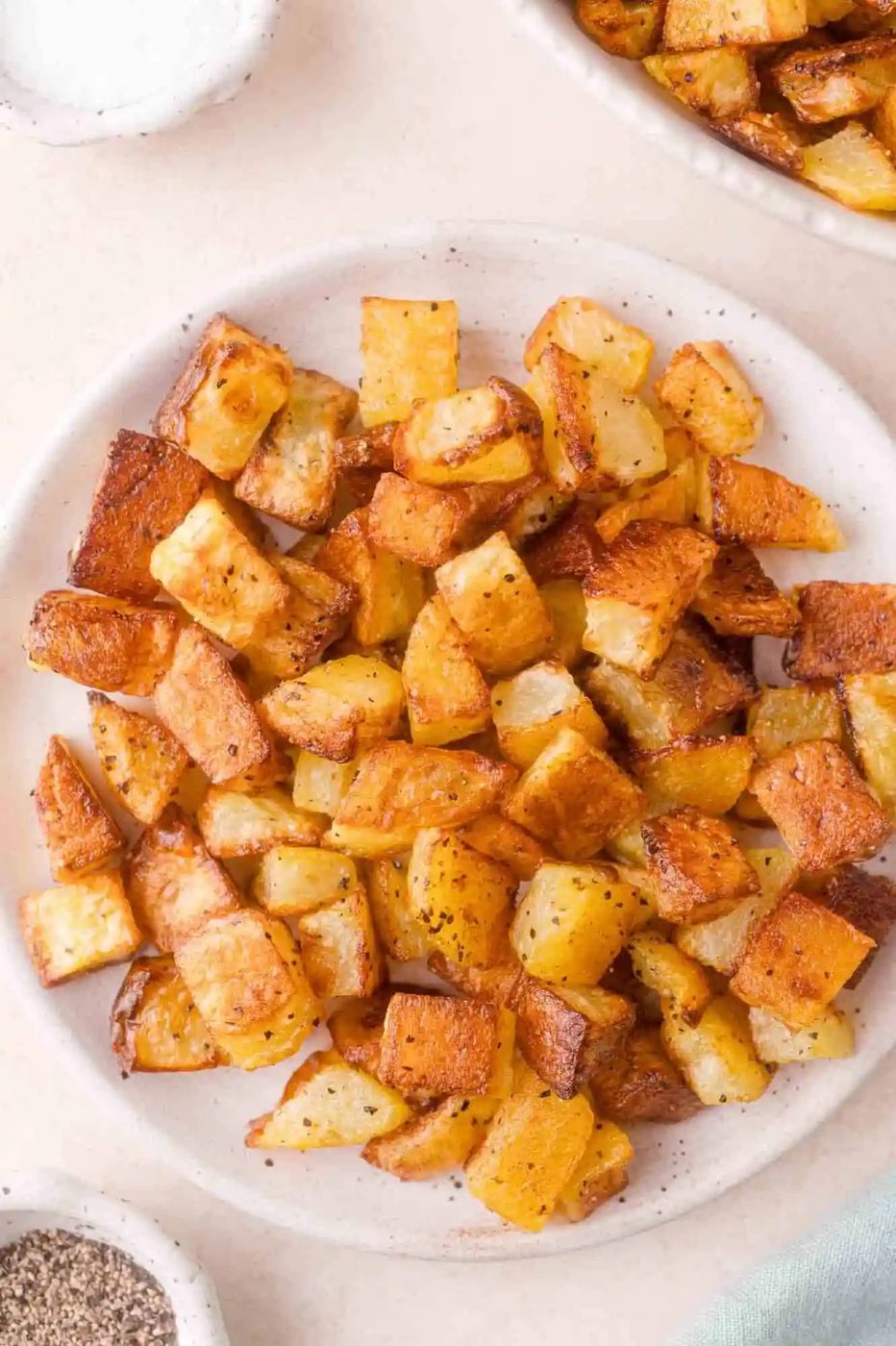 A plate of golden-brown roasted potato cubes, seasoned with pepper, sits on a light-colored surface with small bowls of salt and pepper nearby.