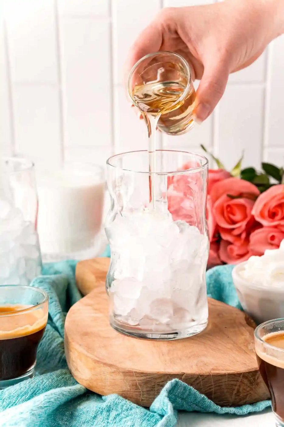 A hand pours liquid from a small glass into a larger glass filled with ice.