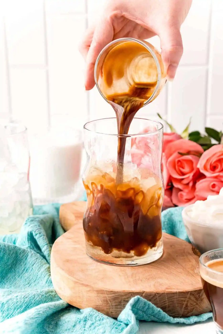 Iced coffee is being poured into a glass on a wooden board, surrounded by ice, milk, and roses, with a teal cloth underneath.