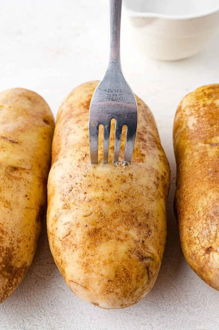 A metal fork piercing the skin of one of three russet potatoes on a white surface. A white bowl is partially visible in the background.