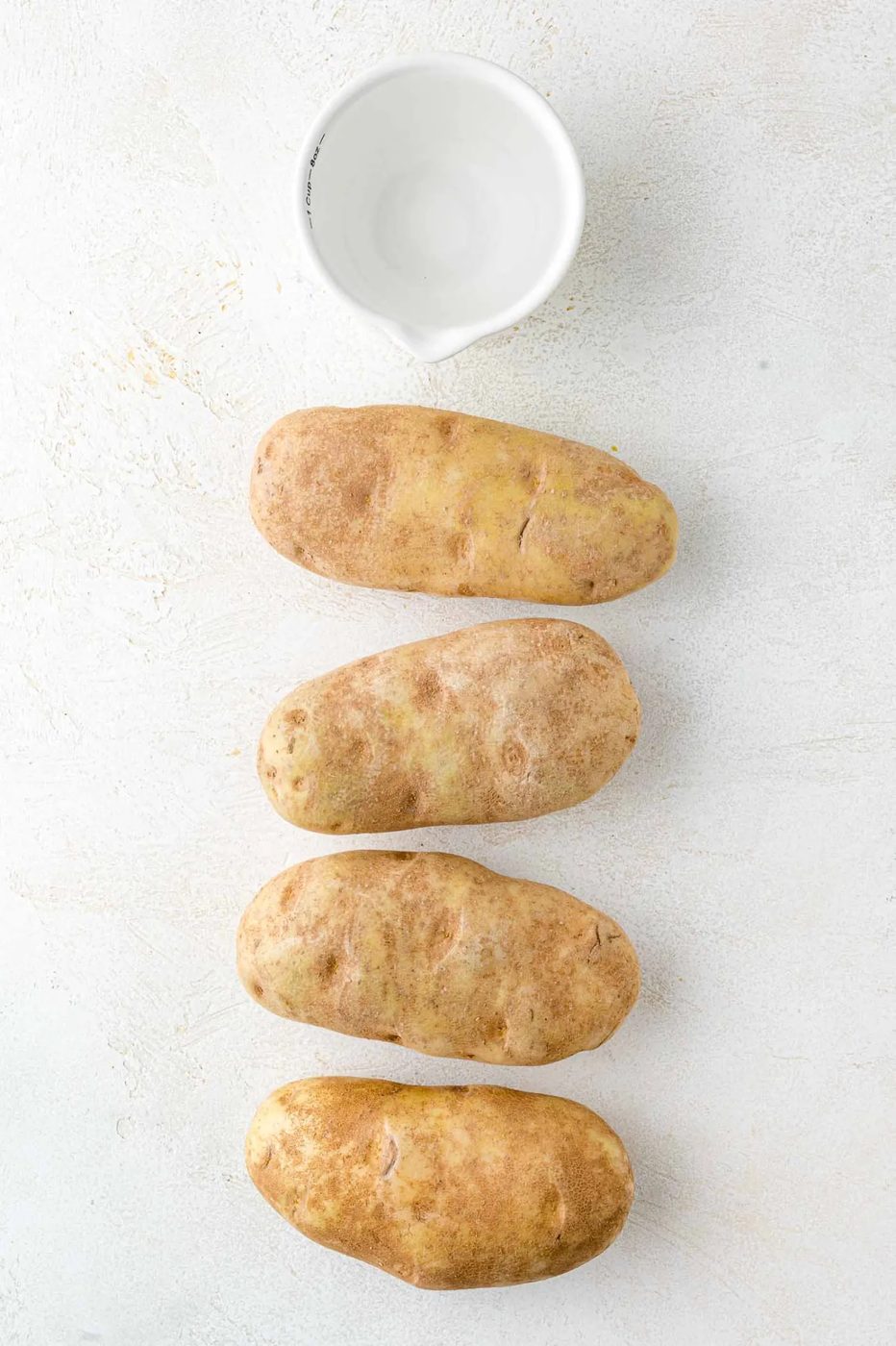 Four potatoes lined vertically on a light surface with a small white bowl above them.