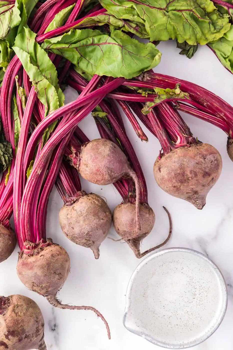 Overhead view of fresh beets with greens attached, and a small measuring cup of water.