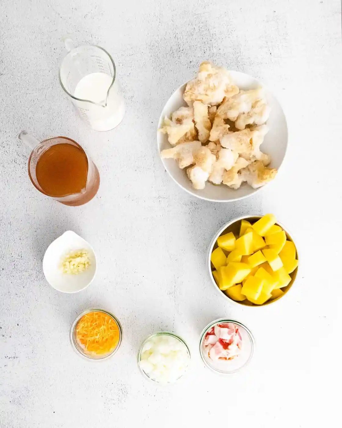 Overhead view of ingredients in bowls: potato, cauliflower, broth, milk, cheese, bacon.
