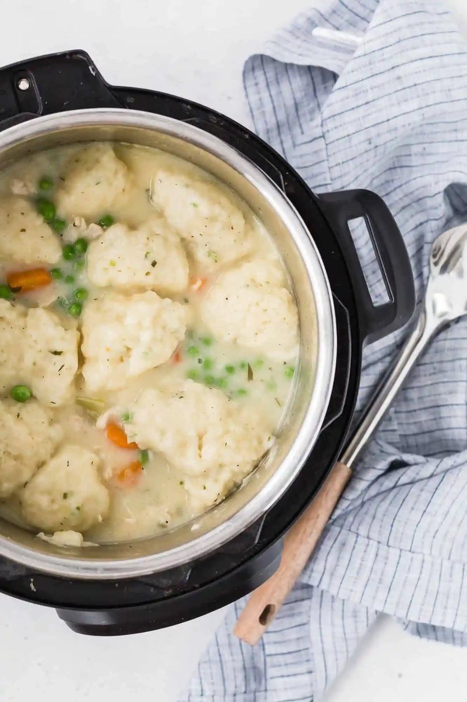 A pot of chicken and dumplings with peas and carrots, next to a cloth and spoon.