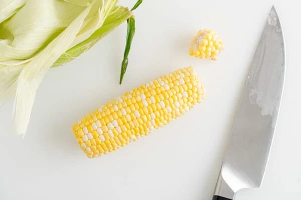 A shucked ear of corn on a countertop with the end trimmed off, next to the green husks and a sharp knife.