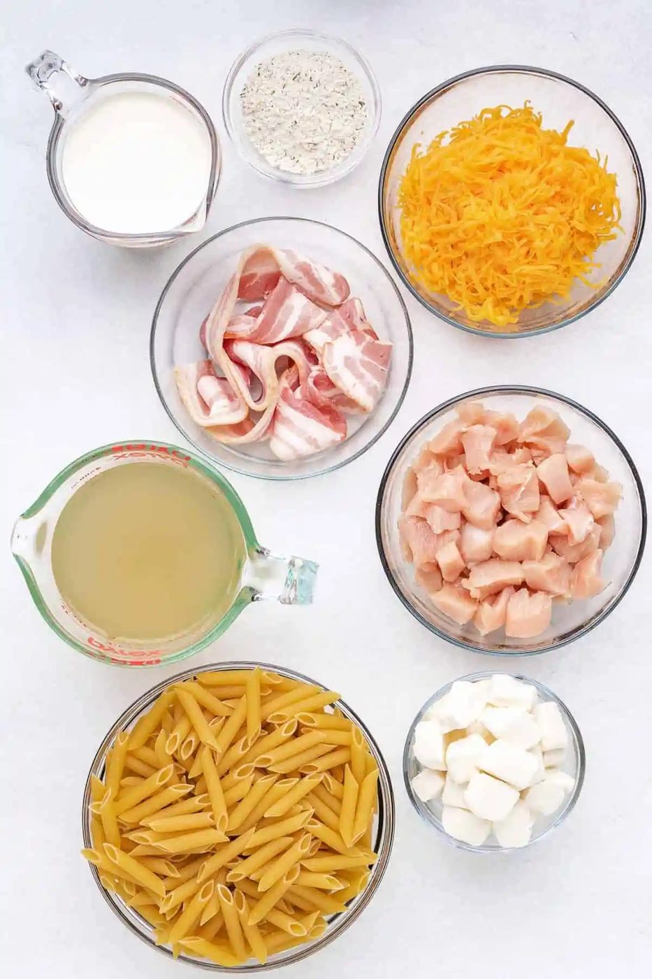 Overhead view of glass bowls of ingredients: pasta, chicken, bacon, cream cheese, cream, ranch, broth.
