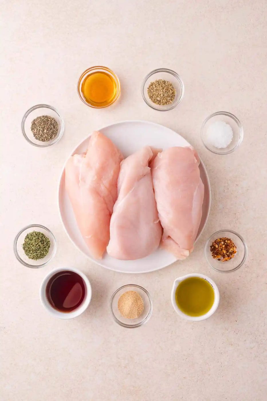 Three raw chicken breasts on a white plate, surrounded by small bowls of various spices, seasonings, and liquids on a light surface.