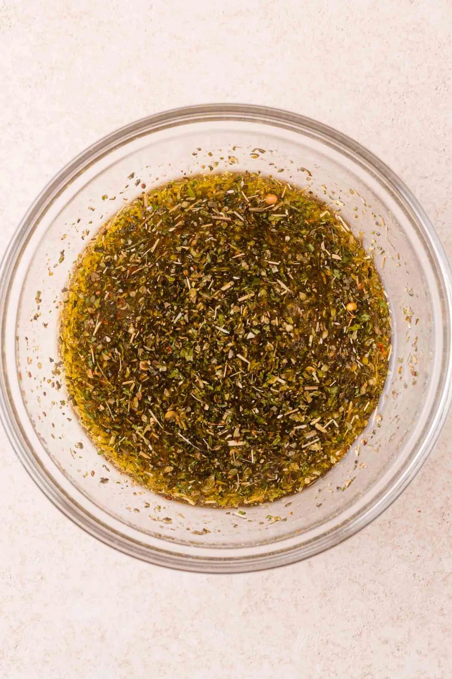A clear glass bowl filled with a mixture of olive oil and various dried herbs and spices on a light-colored surface.