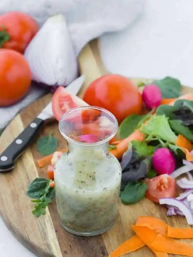 Small glass bottle of from-scratch italian dressing on wooden cutting board surrounded by colorful vegetables.