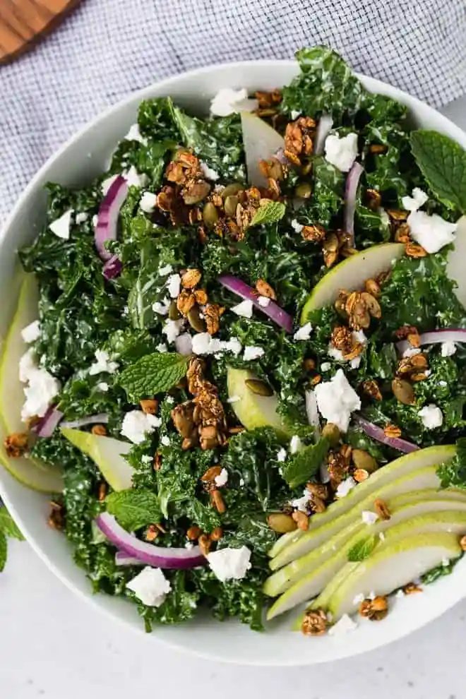 Image of kale and pear salad in a white bowl topped with onions, feta cheese, and savory granola.