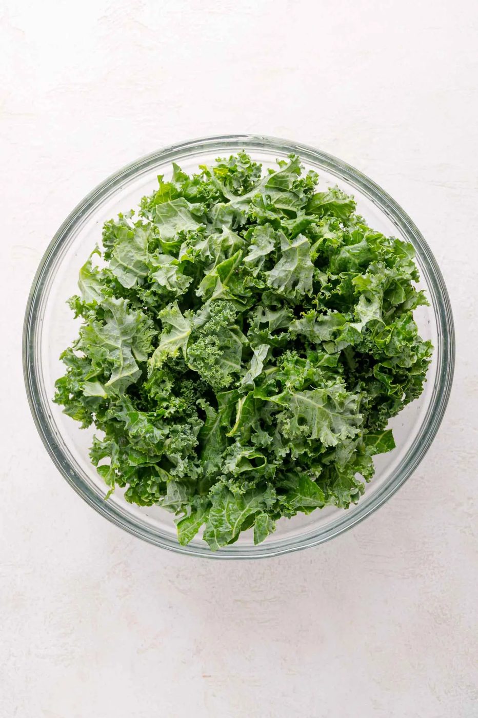 A clear glass bowl filled with fresh, chopped kale leaves, placed on a light-colored surface.