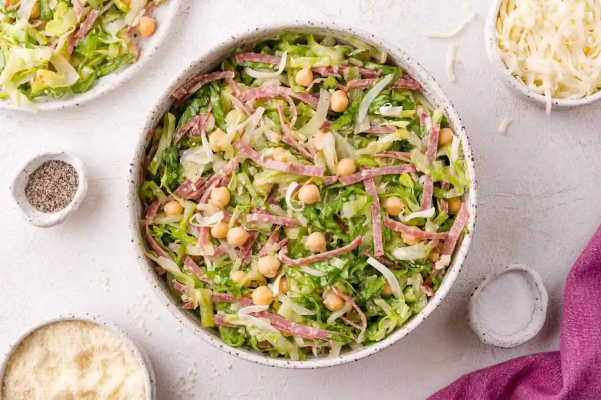 A bowl of chopped la scala salad with shredded lettuce, chickpeas, salami strips, and shredded cheese on a light surface, surrounded by small bowls of ingredients.