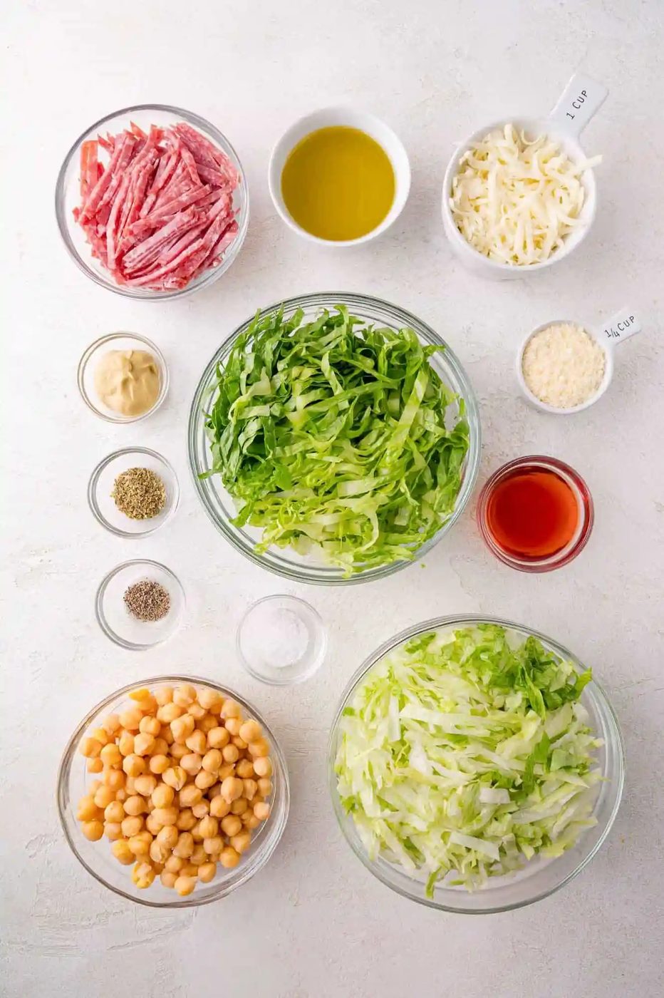 Bowls of shredded lettuce, chickpeas, sliced salami, shredded cheese, grated cheese, olive oil, vinegar, mustard, and seasonings arranged on a white surface.