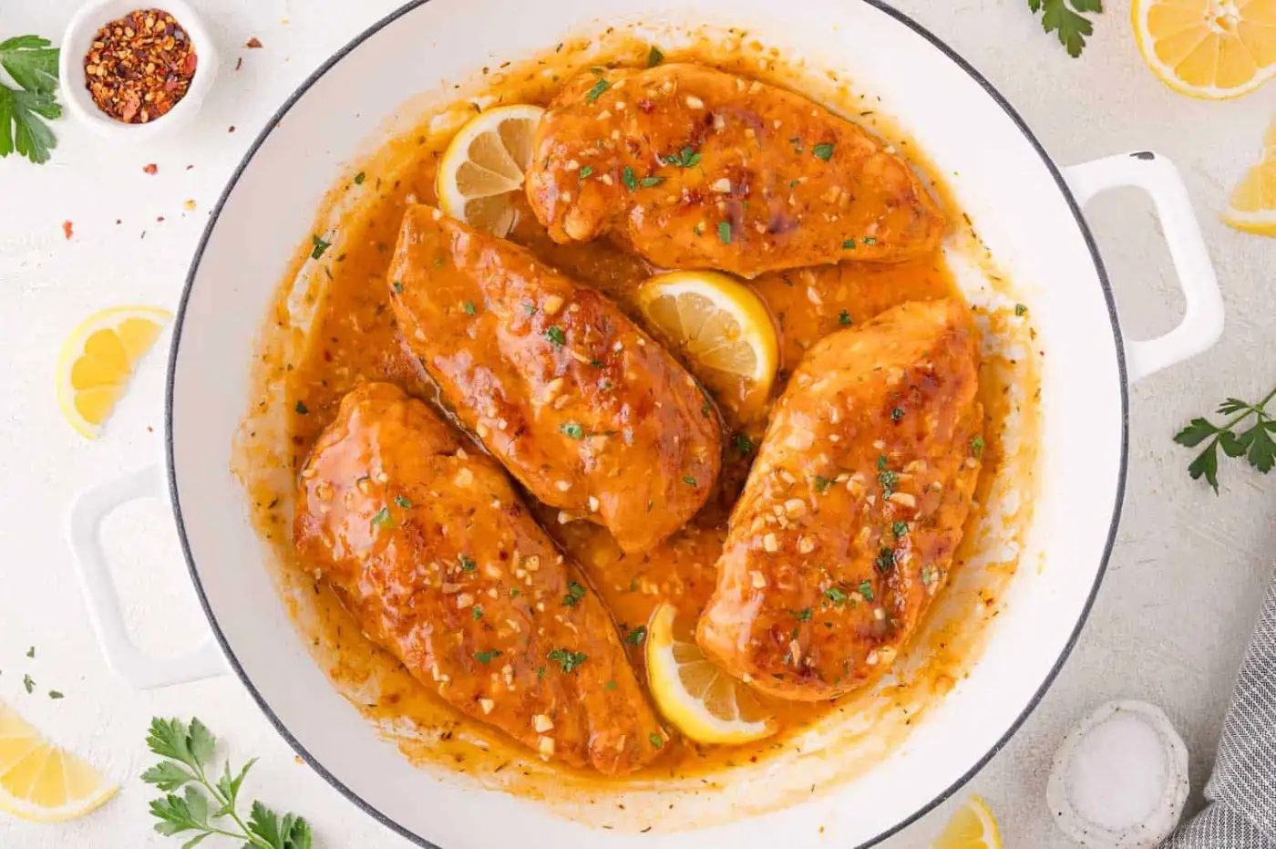 Four cooked chicken breasts in a white pan with a golden lemon-garlic sauce, garnished with lemon slices and chopped herbs.