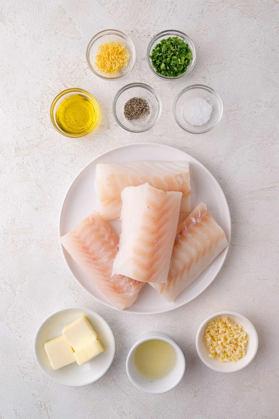 Raw cod fillets on a white plate surrounded by small bowls containing butter, olive oil, lemon juice, minced garlic, lemon zest, parsley, salt, and pepper.