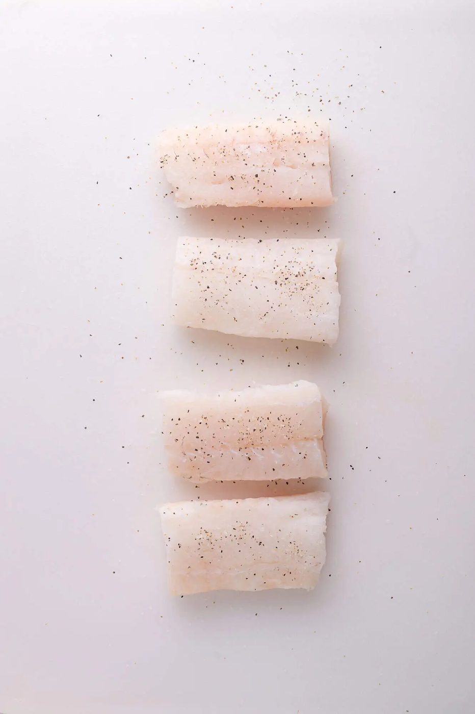 Four raw cod fillets seasoned with salt and pepper are arranged in a vertical line on a light-colored surface.