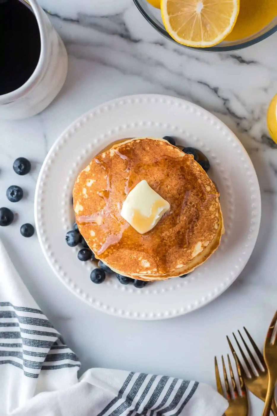 A stack of pancakes topped with butter and syrup on a white plate, surrounded by blueberries. A cup of coffee and lemon slices are nearby, with a striped napkin and gold forks beside the plate.