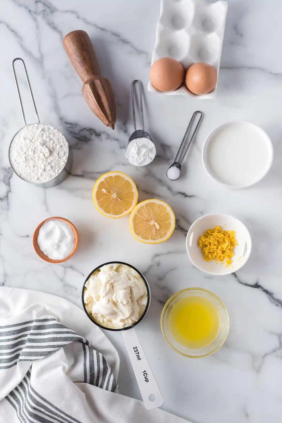 Baking ingredients arranged on a marble surface, including flour, eggs, milk, lemons, butter, and measuring tools.