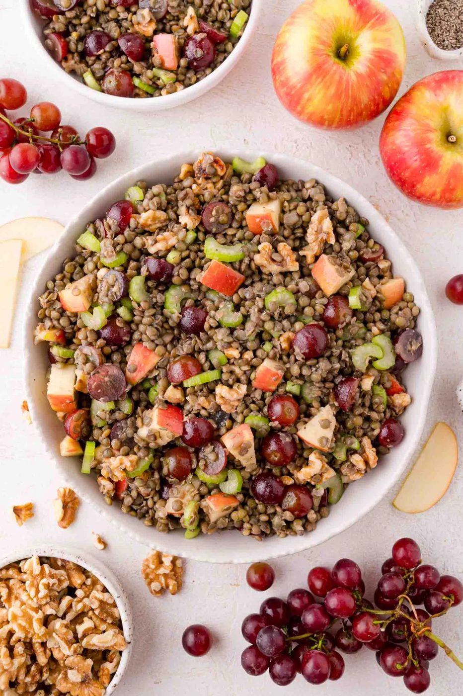 A bowl of lentil salad with grapes, apples, celery, and walnuts, surrounded by fresh apples, grapes, and walnuts on a white surface.