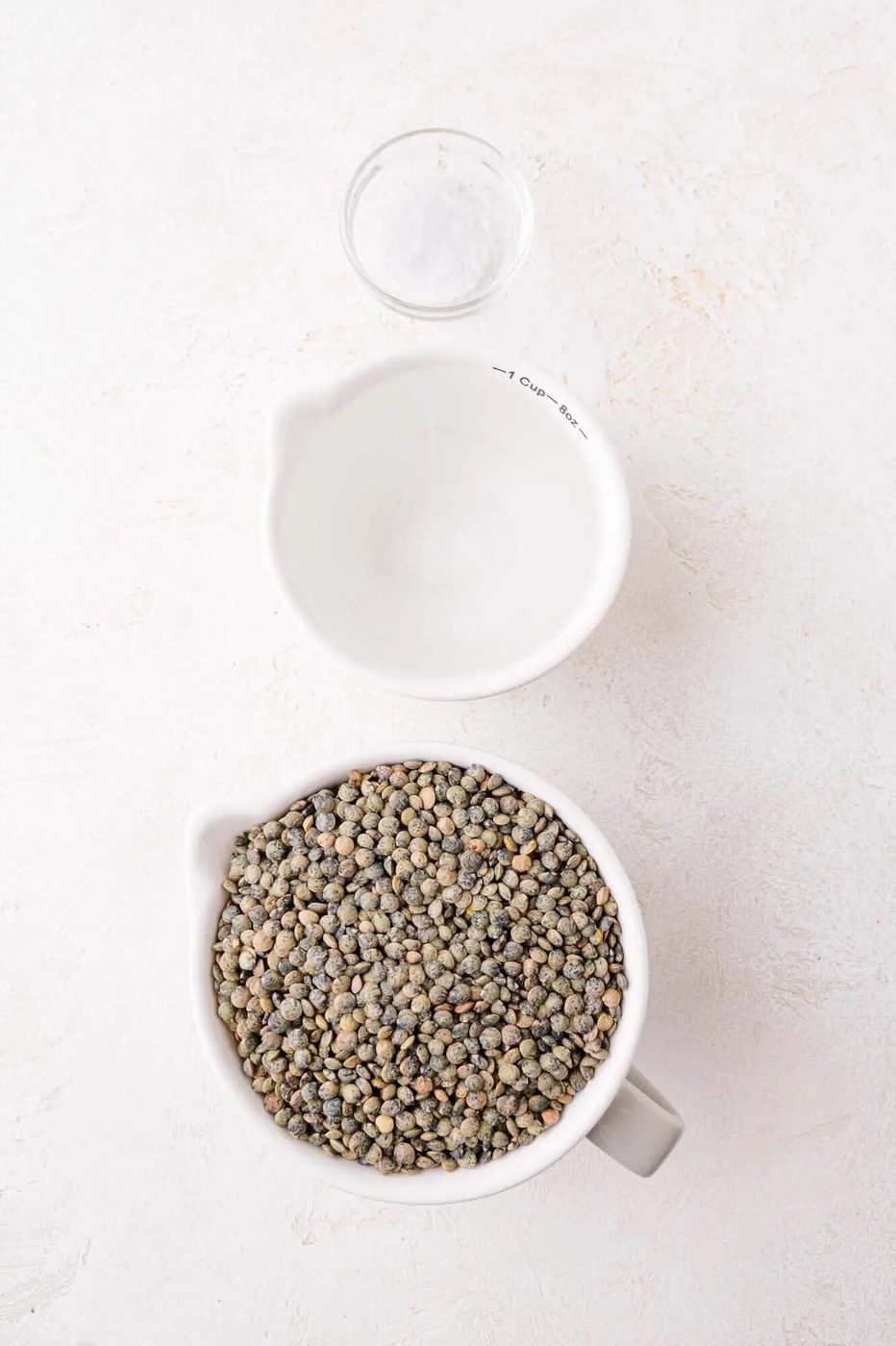 A measuring cup filled with dry lentils, a smaller cup of water, and a small bowl of salt on a light surface.