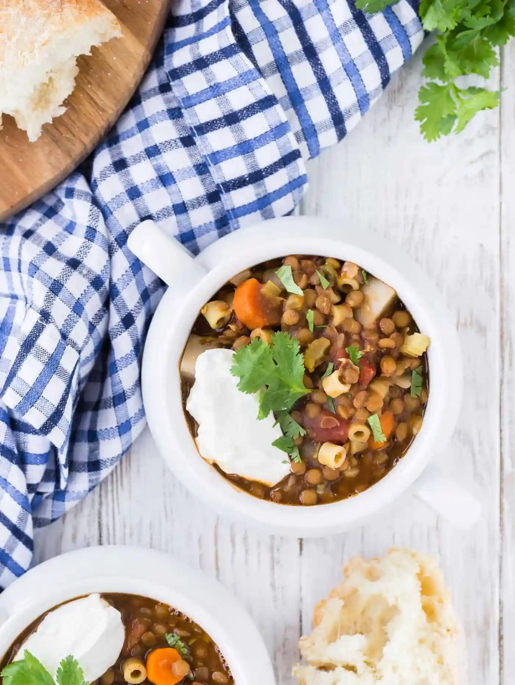Overhead photo of two bowls of lentil soup, one only partially showing in image. Cilantro is also pictured as well as fresh bread that has been torn from the loaf.