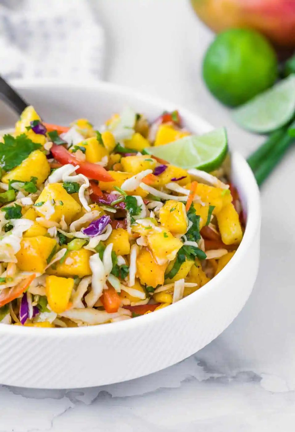 A fresh mango slaw in a white bowl with chopped vegetables, herbs, and lime wedges.