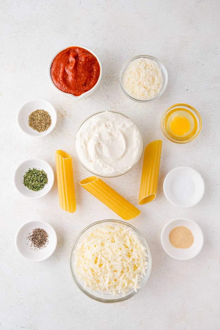 Ingredients for a pasta dish: ricotta, manicotti pasta, tomato sauce, shredded cheese, grated cheese, egg, spices, and seasonings arranged in bowls on a white surface.