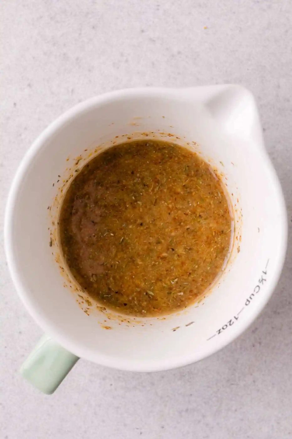 A white measuring cup containing a brown, mustard-based sauce mixture sits on a light gray surface.
