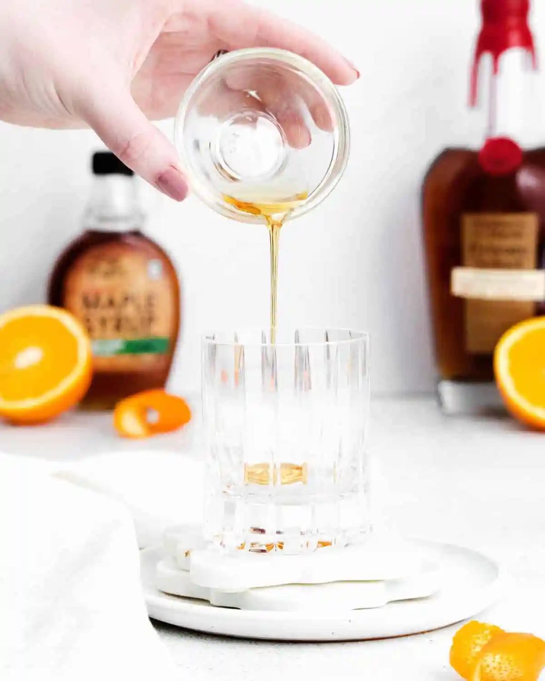 A hand pours syrup from a small glass bowl into an empty glass; orange halves and bottles of syrup and liquor are visible in the background.