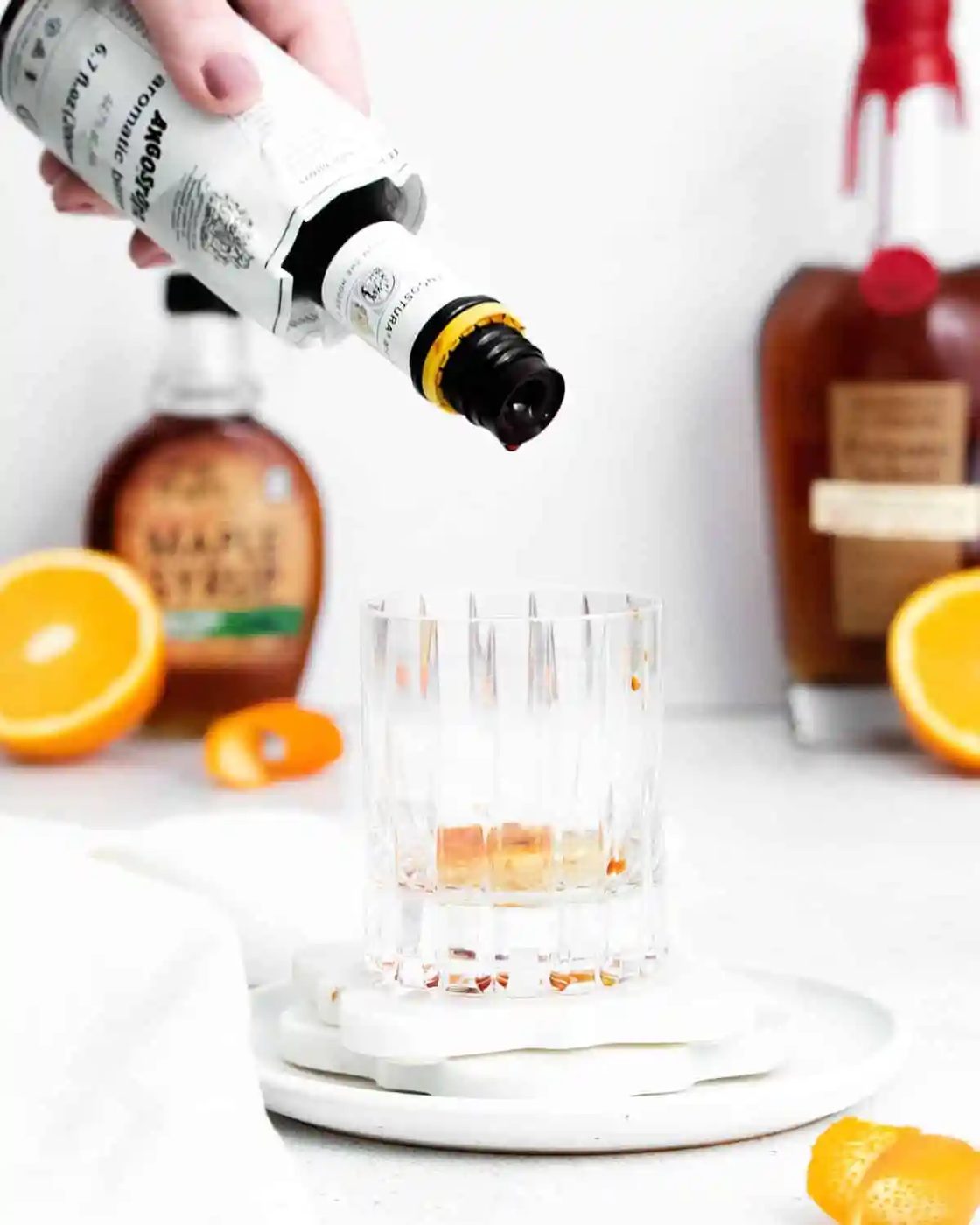 A hand pours bitters into an empty glass on a white surface, with bottles of maple syrup and liquor, and sliced oranges in the background.