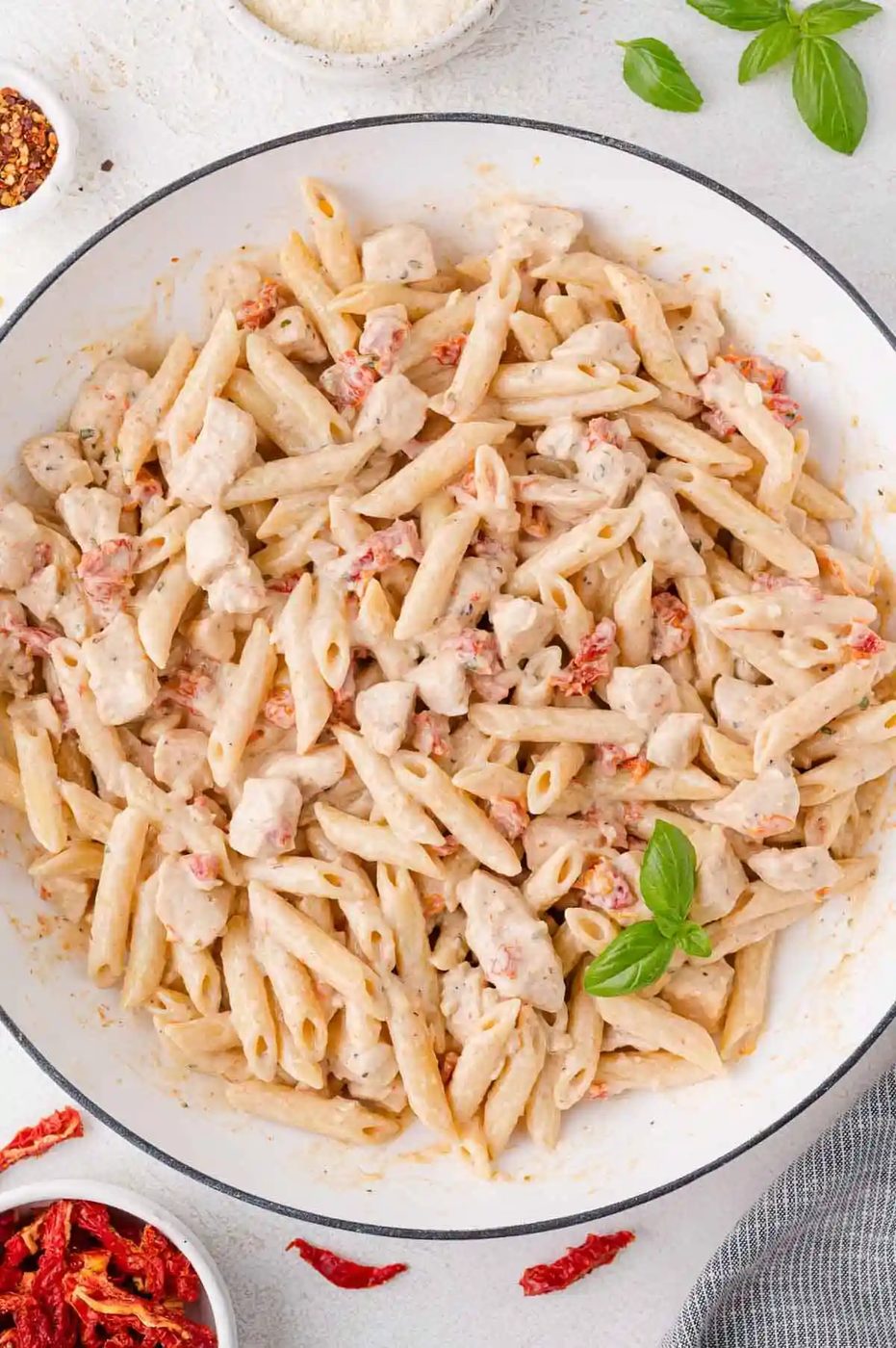 A pan of creamy penne pasta with chicken pieces, sun-dried tomatoes, and a basil garnish.