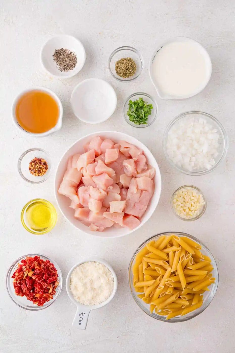 Ingredients arranged on a white surface, including raw chicken pieces, penne pasta, chopped onions, garlic, oil, sun-dried tomatoes, parmesan, cream, spices, and herbs in separate bowls.
