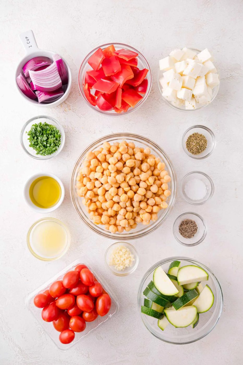 Bowls containing chopped red onion, red bell pepper, halloumi, parsley, olive oil, lemon juice, garlic, grape tomatoes, zucchini, chickpeas, dried herbs, salt, and pepper on a white surface.
